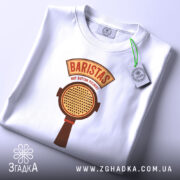 Футболка Бариста білого кольору, складена на поверхні, з видимим принтом. Принт містить текст 'BARISTAS NOT BUTTON PUSHERS' та зображення портафільтра, на футболці підвішена етикетка з зеленим шнурком.