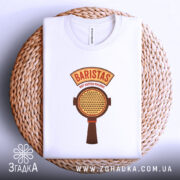 Футболка Бариста білого кольору, складена на плетеній кошику, з видимим принтом. Принт містить текст 'BARISTAS NOT BUTTON PUSHERS' та зображення портафільтра, футболка розташована на світлому фоні.