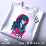 Футболка BIKENT MOM білого кольору, зображена з верхнього ракурсу, складена на поверхні, фон світлий, принт з дівчиною в сонцезахисних окулярах та текстом 'BIKENT MOM' в яскравих кольорах, на футболці є етикетка.