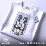 Футболка бойові гуси, біла, зображена зверху, на столі, фон світло-сірий, принт з двома гусами в військових шоломах і з автоматами, на футболці є етикетка з зеленим шнурком.