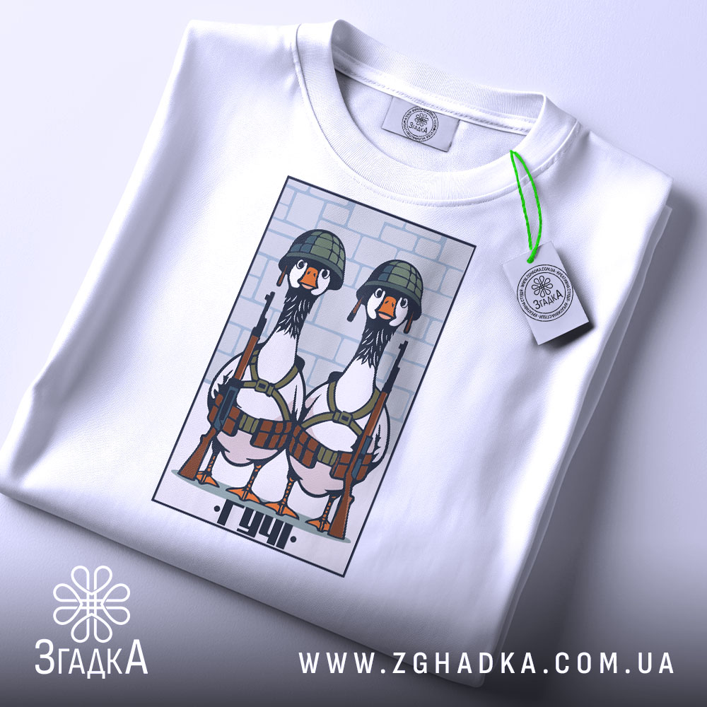 Футболка бойові гуси, біла, зображена зверху, на столі, фон світло-сірий, принт з двома гусами в військових шоломах і з автоматами, на футболці є етикетка з зеленим шнурком.