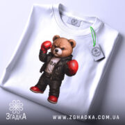 Футболка боксерський ведмідь, біла, складена на столі, фон світло-сірий, принт ведмедя в боксерських рукавичках та шкіряному жакеті, з червоними черевиками, з етикеткою на рукаві.
