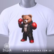 Футболка боксерський ведмідь, біла, складена на столі, фон світло-сірий, принт ведмедя в боксерських рукавичках та шкіряному жакеті, з червоними черевиками, з етикеткою на рукаві.