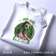 Футболка Brr brr Patapim білого кольору, складена на поверхні, фон світлий, принт з персонажем з великими руками та ногами на зеленому фоні, прикріплена етикетка з зеленим шнурком.