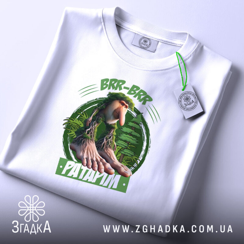 Футболка Brr brr Patapim білого кольору, складена на поверхні, фон світлий, принт з персонажем з великими руками та ногами на зеленому фоні, прикріплена етикетка з зеленим шнурком.