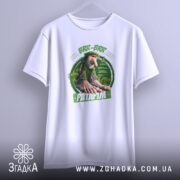 Футболка Brr brr Patapim білого кольору, підвішена на вішалці, фон світлий, принт з персонажем з великими руками та ногами на зеленому фоні, текст на принті виділений зеленим кольором.