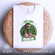 Футболка Brr brr Patapim білого кольору, складена на плетеній основі, фон світлий, принт з персонажем з великими руками та ногами на зеленому фоні, текст на принті виділений зеленим кольором.