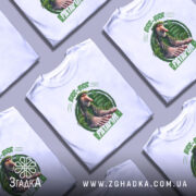 Футболка Brr brr Patapim білого кольору, зображена на манекені з блискучою поверхнею, фон нейтральний, принт з персонажем з великими руками та ногами на зеленому фоні з рослинними елементами.