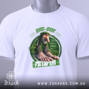 Футболка Brr brr Patapim білого кольору, складена на поверхні, фон світлий, принт з персонажем з великими руками та ногами на зеленому фоні, прикріплена етикетка з зеленим шнурком.