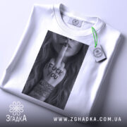 Футболка бунтарський арт, біла, складена на столі, фон світло-сірий, принт у чорному та білому кольорах з дівчиною, що показує середній палець, на футболці є етикетка з логотипом.