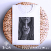 Футболка бунтарський арт, біла, складена на плетеній кошику, фон світло-сірий, принт у чорному та білому кольорах з дівчиною, що показує середній палець, на футболці видно етикетку.