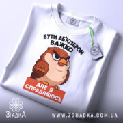 Футболка бути абʼюзером білого кольору, зображена зверху, на плоскій поверхні, фон світлий, принт з карикатурним птахом, текст чорного кольору зверху і оранжевого знизу, на футболці є зелений ярлик.