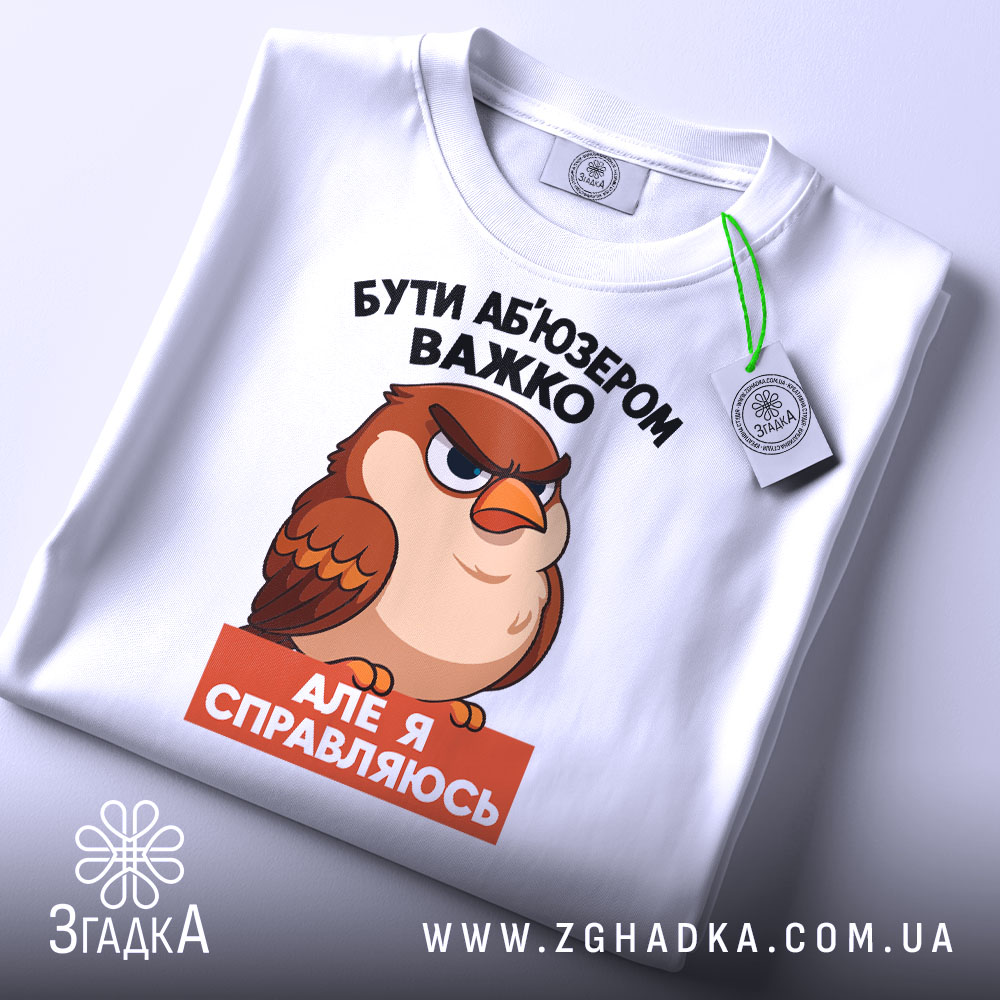 Футболка бути абʼюзером білого кольору, зображена зверху, на плоскій поверхні, фон світлий, принт з карикатурним птахом, текст чорного кольору зверху і оранжевого знизу, на футболці є зелений ярлик.