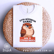 Футболка бути абʼюзером білого кольору, зображена зверху на плоскій поверхні, фон білий, принт з карикатурним птахом, текст чорного кольору зверху і оранжевого знизу, футболка лежить на плетеній основі.