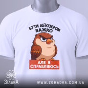 Футболка бути абʼюзером білого кольору, зображена зверху, на плоскій поверхні, фон світлий, принт з карикатурним птахом, текст чорного кольору зверху і оранжевого знизу, на футболці є зелений ярлик.