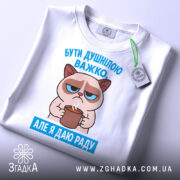 Футболка бути душнілою, білого кольору, зображена в горизонтальному положенні, фон світло-сірий, принт з котом, що тримає чашку, з текстом 'БУТИ ДУШНІЛЮ ВАЖКО. АЛЕ Я ДАЮ РАДУ' у блакитному кольорі, на футболці є етикетка з зеленим шнурком.