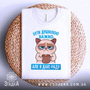 Футболка бути душнілою, білого кольору, зображена в горизонтальному положенні на плетеному кошику, фон світло-сірий, принт з котом, що тримає чашку, з текстом 'БУТИ ДУШНІЛЮ ВАЖКО. АЛЕ Я ДАЮ РАДУ' у блакитному кольорі.