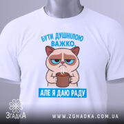 Футболка бути душнілою, білого кольору, зображена в горизонтальному положенні, фон світло-сірий, принт з котом, що тримає чашку, з текстом 'БУТИ ДУШНІЛЮ ВАЖКО. АЛЕ Я ДАЮ РАДУ' у блакитному кольорі, на футболці є етикетка з зеленим шнурком.