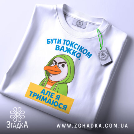 Футболка бути токсіком, біла, зображена зверху на плоскій поверхні, фон світло-сірий, принт з персонажем у зеленому капюшоні та текстом жовтого кольору, на комірці видна етикетка.
