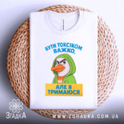 Футболка бути токсіком, біла, зображена на плоскій поверхні, фон світло-сірий, принт з персонажем у зеленому капюшоні та текстом жовтого кольору, розташована на плетеній кошику.