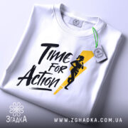 Футболка час для дії, біла, складена на столі, з видимим етикеткою з зеленим шнурком, фон світло-сірий, принт чорного кольору з жовтою блискавкою та силуетом жінки, що біжить.