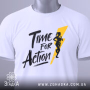 Футболка час для дії, біла, складена на столі, з видимим етикеткою з зеленим шнурком, фон світло-сірий, принт чорного кольору з жовтою блискавкою та силуетом жінки, що біжить.