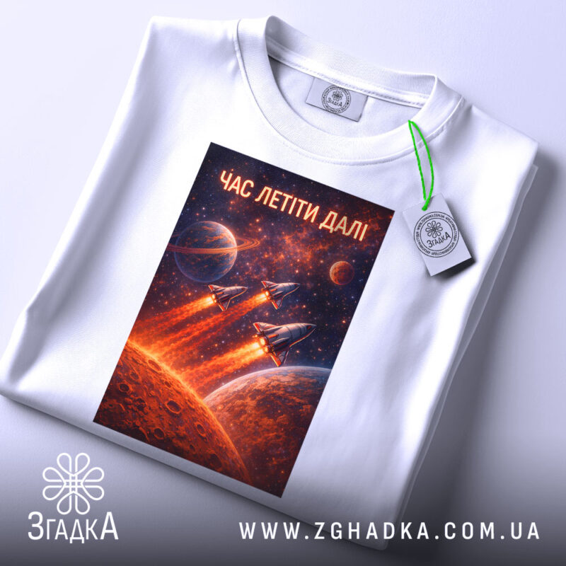 Футболка час летіти далі. Біла футболка, складена на столі, з принтом космічної тематики, на якому зображені ракети, що злітають, на фоні червоної планети та зірок. Вгорі напис 'ЧАС ЛЕТІТИ ДАЛІ', а також видно етикетку з зеленим шнурком.