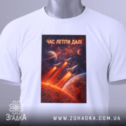 Футболка час летіти далі. Біла футболка, складена на столі, з принтом космічної тематики, на якому зображені ракети, що злітають, на фоні червоної планети та зірок. Вгорі напис 'ЧАС ЛЕТІТИ ДАЛІ', а також видно етикетку з зеленим шнурком.