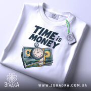 Футболка час це гроші, складена біла футболка з принтом, видно етикетку з зеленим шнурком, фон світло-сірий, принт яскравий, зображення годинника та купюр.