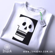 Футболка череп білого кольору, складена на столі, фон світло-сірий, принт чорного кольору з малюнком черепа, що має великі очі та зуби, прикріплена етикетка з зеленим шнурком.