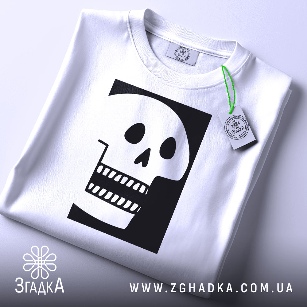 Футболка череп білого кольору, складена на столі, фон світло-сірий, принт чорного кольору з малюнком черепа, що має великі очі та зуби, прикріплена етикетка з зеленим шнурком.