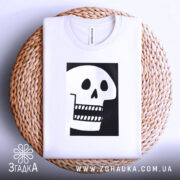 Футболка череп білого кольору, складена на плетеній кошику, фон світло-сірий, принт чорного кольору з малюнком черепа, що має великі очі та зуби.