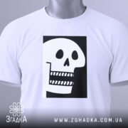 Футболка череп білого кольору, складена на столі, фон світло-сірий, принт чорного кольору з малюнком черепа, що має великі очі та зуби, прикріплена етикетка з зеленим шнурком.