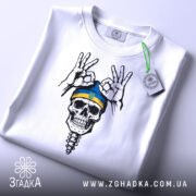 Футболка череп воїн, біла, складена на столі, зображена з верхнього ракурсу, фон світло-сірий, принт з черепом у блакитній шапці з жовтою смужкою та жестами рук, на футболці є етикетка.