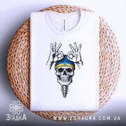 Футболка череп воїн, біла, складена на плетеному кошику, зображена з верхнього ракурсу, фон світло-сірий, принт з черепом у блакитній шапці з жовтою смужкою та жестами рук.