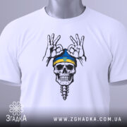 Футболка череп воїн, біла, складена на столі, зображена з верхнього ракурсу, фон світло-сірий, принт з черепом у блакитній шапці з жовтою смужкою та жестами рук, на футболці є етикетка.