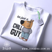 Футболка chill guy стиль, біла, зображена зверху, складена, фон світло-сірий, принт з мультяшним ведмедем у джинсах та светрі, текст 'im just a CHILL GUY', на комірці видна етикетка.