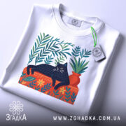 Футболка Чорний кіт спокою білого кольору, зображена зверху, складена на поверхні, фон світло-сірий. Принт з чорним котом, що лежить на червоній підставці, оточений зеленими рослинами та квітами, з етикеткою на рукаві.