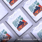 Футболка Чорний кіт спокою білого кольору, зображена з фронтального ракурсу на манекені, фон світло-сірий. Принт з чорним котом, що лежить на червоній підставці, оточений зеленими рослинами та квітами.