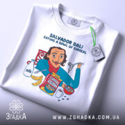 Футболка Далі арт принт, біла, зображена зверху, фон світло-сірий, принт з ілюстрацією Сальвадора Далі, який їсть з миски, з деталями молока, бананів і коробки з написом 'Surreal', на комірці видна етикетка.