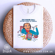 Футболка Далі арт принт, біла, складена на плетеній основі, фон світло-сірий, принт з ілюстрацією Сальвадора Далі, який їсть з миски, з деталями молока, бананів і коробки з написом 'Surreal'.