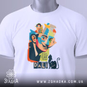 Футболка Далі арт, біла, зображена на столі, фон світло-сірий, принт з обличчям художника, кольорові деталі, текст "I don't do drugs I AM drugs", на комірці етикетка з зеленим шнурком.