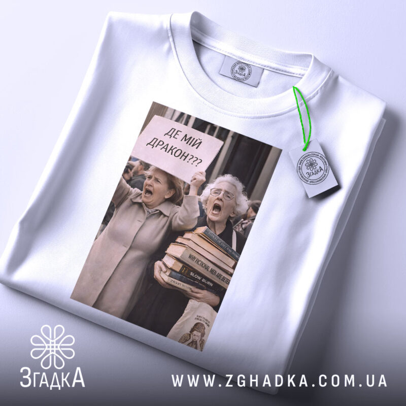Футболка де мій дракон, біла, зображена зверху, на фоні світло-сірого кольору, принт з двома жінками, які вигукують з плакатом 'ДЕ МІЙ ДРАКОН???', на футболці є етикетка з зеленим шнурком.