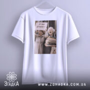 Футболка де мій дракон, біла, зображена з переднього ракурсу, на фоні світло-сірого кольору, принт з двома жінками, які тримають плакат з написом 'ДЕ МІЙ ДРАКОН???' та купою книг, футболка висить на вішалці.