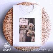 Футболка де мій дракон, біла, зображена зверху на плетеній кошику, на фоні світло-сірого кольору, принт з двома жінками, які тримають плакат з написом 'ДЕ МІЙ ДРАКОН???' та купою книг.