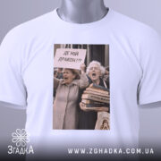 Футболка де мій дракон, біла, зображена зверху, на фоні світло-сірого кольору, принт з двома жінками, які вигукують з плакатом 'ДЕ МІЙ ДРАКОН???', на футболці є етикетка з зеленим шнурком.