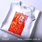 Футболка демон червоний, біла футболка на плоскій поверхні, ракурс зверху, фон світло-блакитний, принт з червоним демоном та черепами, з етикеткою на зеленому шнурку.