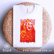 Футболка демон червоний, біла футболка на плоскій поверхні, ракурс зверху, фон світло-блакитний, принт з червоним демоном та черепами, розташована на плетеній основі.