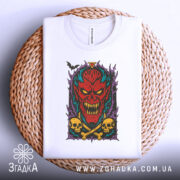 Футболка демонічний яскравий образ, біла, з верхнього ракурсу, на плетеній кошику, фон світло-сірий, принт з червоним демоном, жовтими черепами та чорними кістками, оточений фіолетовими елементами.