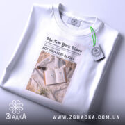 Футболка дівчата читають книги, біла, з верхнього ракурсу, складена на поверхні, фон світлий, принт у стилі газети з текстом 'HOT GIRLS READ BOOKS' та зображенням книги з навушниками.